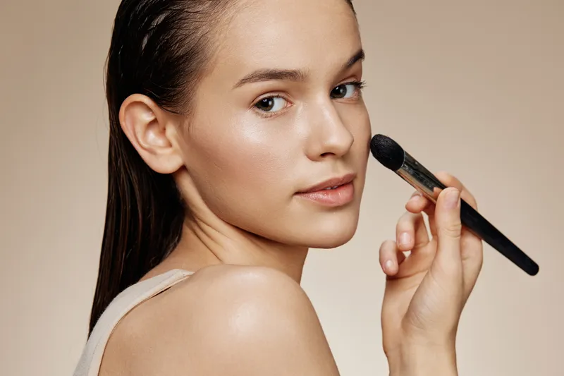 Young model applying makeup with a brush, showcasing a natural beauty look and smooth skin against a soft, neutral backdrop Ideal for beauty and skincare concepts