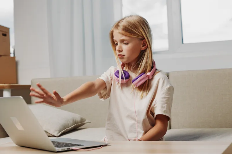 Blond girl with headphones sitting on sofa uses laptop, showing stop gesture with hand. Concept for online safety, digital boundaries, and child screen time management.