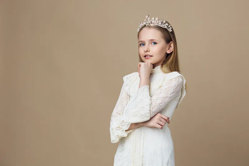 Young girl wearing a delicate white dress with a crown, exuding grace and elegance against a soft beige background, showcasing beauty and innocence