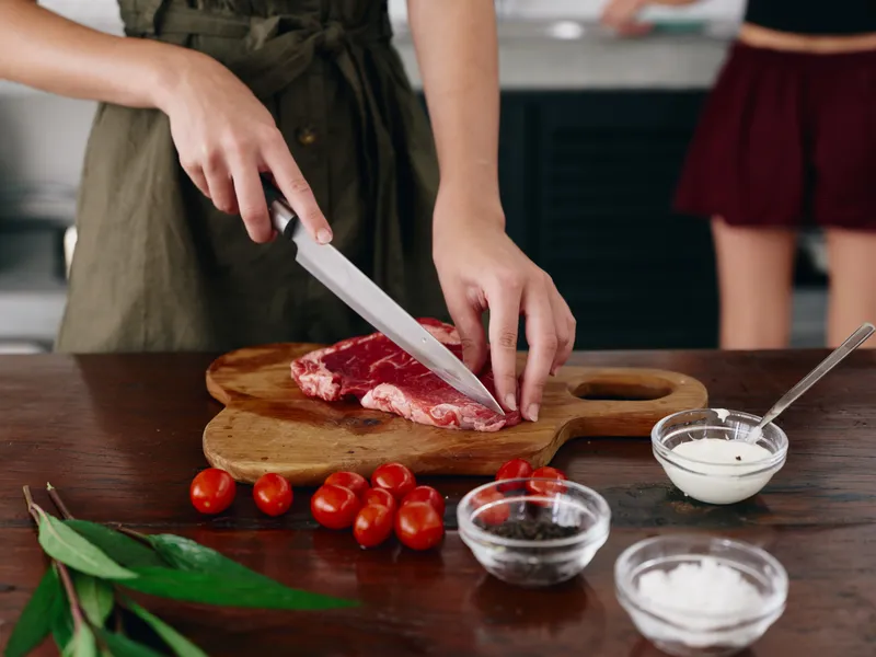 Женщина с ножом в руке режет сырое мясо для жарки на кухне