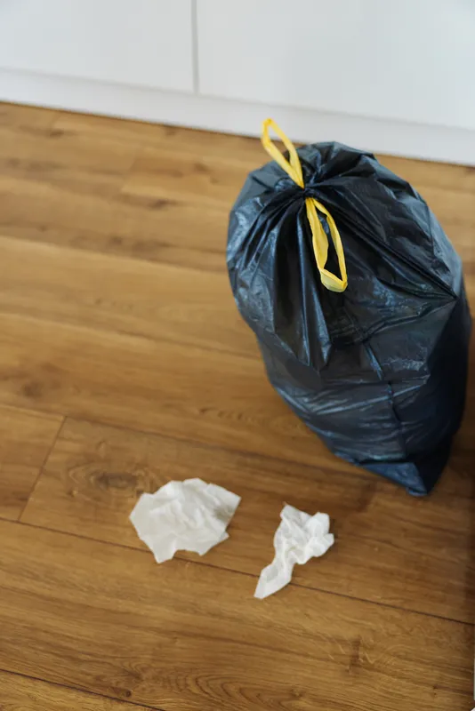 Garbage trash bag waste rubbish floor: black plastic sack tied with yellow string on hardwood, crumpled tissues nearby, domestic disposal and household cleaning scene for tidy up.