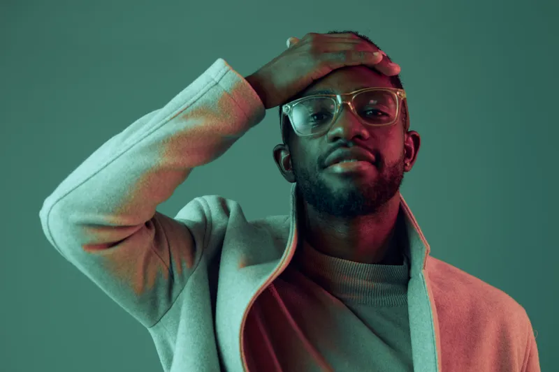 Stylish young Black man in glasses posing with a hand on his forehead, against a vibrant green and pink background, showcasing a modern fashion concept