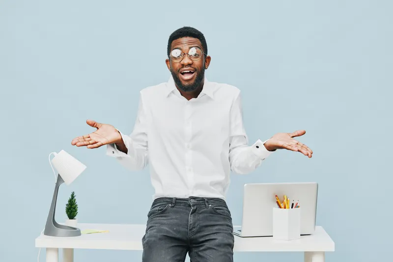 Confused man with glasses, unsure expression, gesturing with palms up, dressed in white shirt, standing behind modern white desk with laptop and office supplies, isolated on light blue background, stu