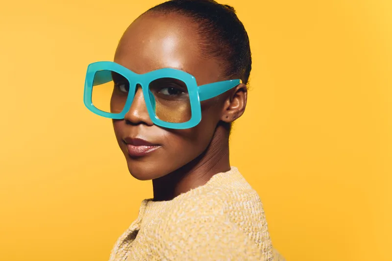 Stylish African woman with dark skin wearing large blue sunglasses and yellow sweater poses confidently against vibrant yellow background. Concept of fashion, beauty, and modern style.