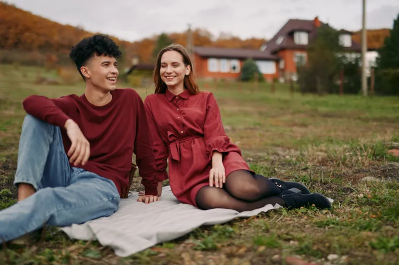 A candid outdoor portrait capturing authentic connection between two friends sitting on a blanket in an autumn field, with relaxed smiles and natural, spontaneous mood.