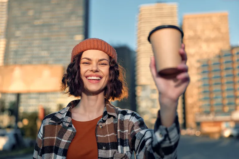 Woman holding coffee in city street portrait, smile and candid expression during golden hour glow, authenticity and mindful living with emotional storytelling urban lifestyle vibe.