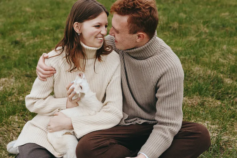 Lifestyle couple outdoors goat kid affection nature warmth cozy harmony, a candid moment with a smiling pair in knitwear, gentle animal presence, relaxed atmosphere, natural colors