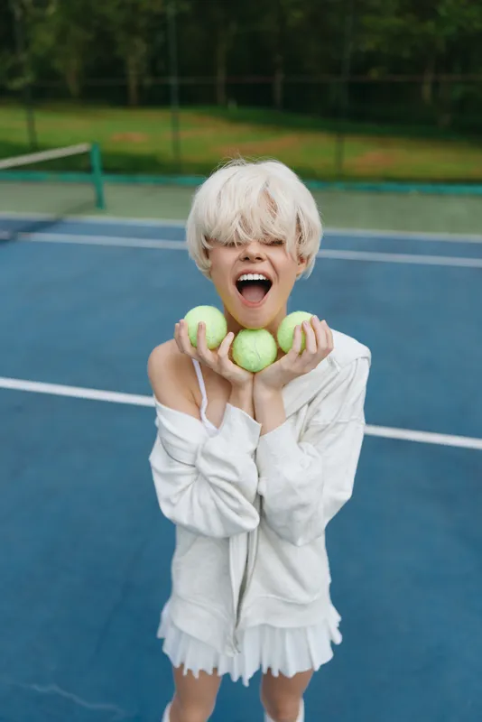 Happy young woman with short blond hair holding tennis balls on a blue court, dressed casually in a white sweater, expressing joy and enthusiasm for sports activities