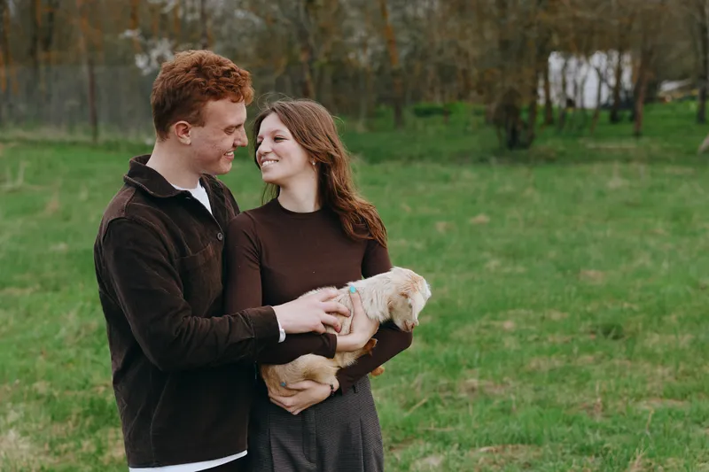 Joyful couple outdoors, holding a young goat kid, warm smiles, gentle touch, autumn light, casual sweaters, grassy meadow, candid lifestyle moment, natural colors, countryside romance