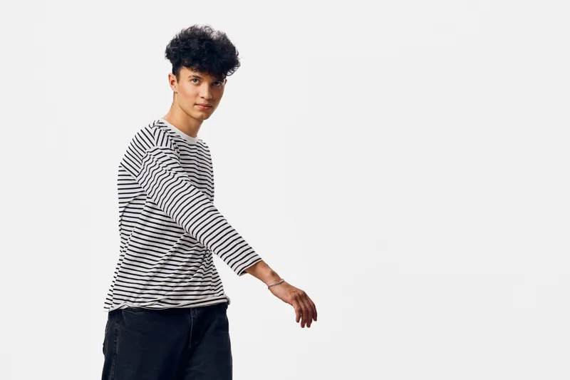young man wearing striped long sleeve shirt and black pants, standing with relaxed posture against white background, casual style portrait