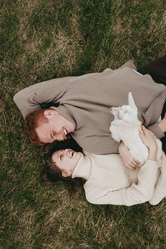 A couple lies on grass, embracing a gentle goat kid between them, capturing a warm lifestyle moment in nature. Cozy sweaters, soft light, and shared joy convey bonding.