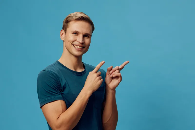 Smiling young man with short hair wearing casual blue t-shirt pointing with both hands, cheerful expression, blue background, studio portrait, lifestyle concept, happiness, positivity, young adult.