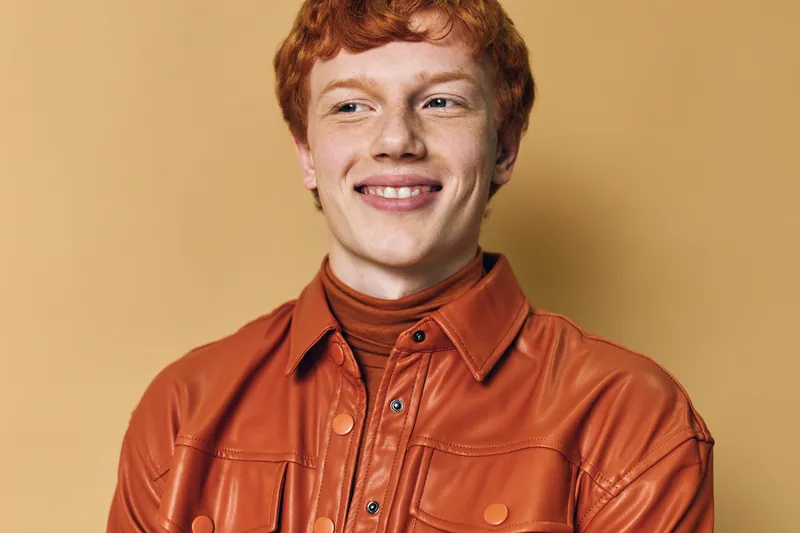 Young man with red hair wearing brown leather jacket and turtleneck smiling with head turned slightly on beige background, casual style and confident expression in portrait.