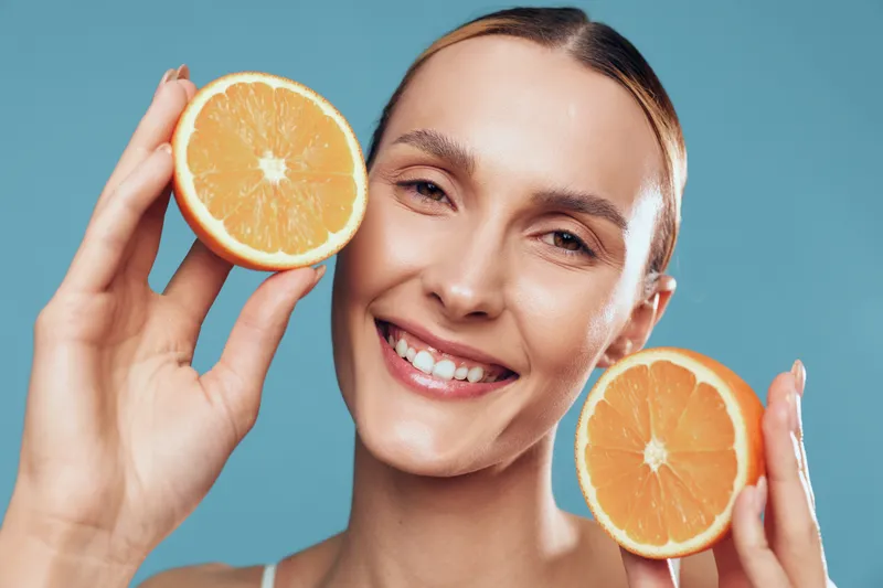 Smiling woman holding two orange halves near face, fresh citrus fruit, healthy skin, natural makeup, bright background, cheerful expression, vitamin C concept.