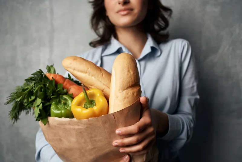 Улыбающаяся женщина с пакетом продуктов, в котором лежат овощи, совершает покупки в супермаркете