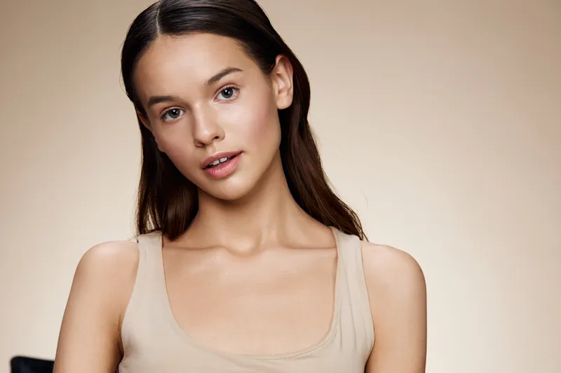 Natural beauty portrait of a young woman with long hair, wearing a simple beige top, displaying a soft expression against a neutral background
