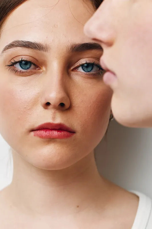 Portrait of young woman with blue eyes and red lips looking at camera with blurred man profile close to her face in minimalistic background.
