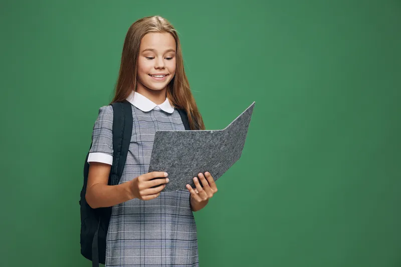 Schoolgirl with backpack reading document or folder standing against green background for education, student, learning and childhood. Girl smiling with school uniform on.