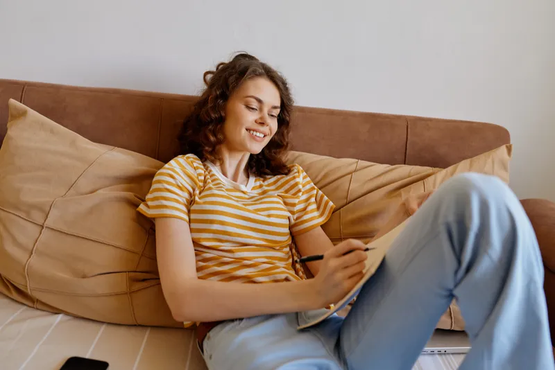 Smiling woman with striped shirt relaxing on a couch while using a tablet. Bright, cozy living room creates a warm home vibe and comfortable personal time, with calm mood.