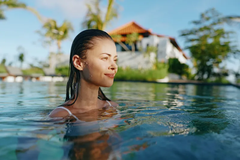Relaxed woman swims in a sunlit tropical pool, enjoying calm water and a warm breeze, with a resort villa and palm trees in the background. She smiles softly, savoring vacation calm.