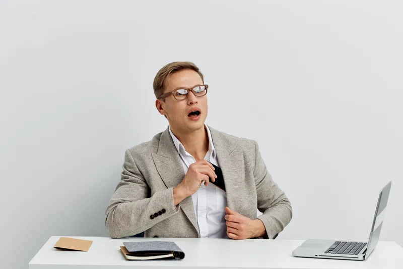 Frustrated man in business attire sitting at desk with laptop, holding tie and showing tired expression, in modern office environment. Workplace stress concept, professional fatigue.
