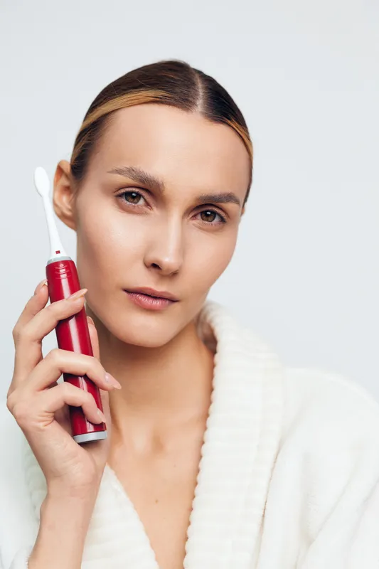 A young woman with clear skin holds a red electric toothbrush near her face wearing a white bathrobe. The close-up portrait emphasizes dental hygiene and personal care concept.