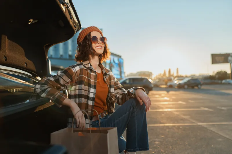 Woman car shopping trunk smile sunglasses golden parking scene with shopping bags and sunlight, candid lifestyle portrait conveying authenticity, mindful living and warm emotional storytelling.