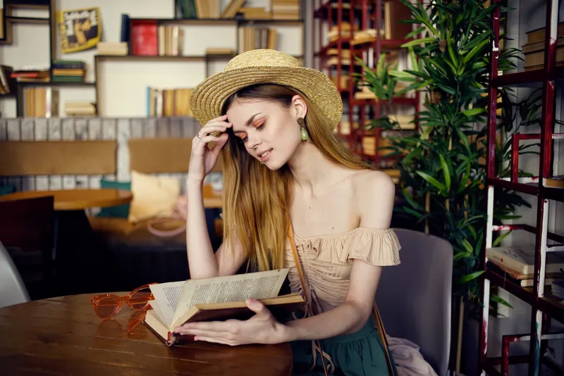 Симпатичная женщина в шляпе сидит за столом с книгами на заднем плане
