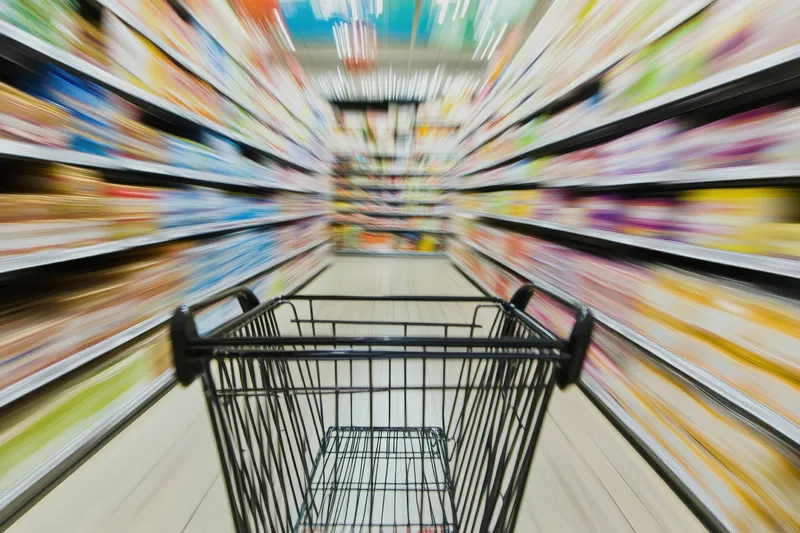 Пустая тележка для покупок в проходе продуктового магазина, где товары на полках размыты в движении, что создает динамичную атмосферу и опыт совершения покупок