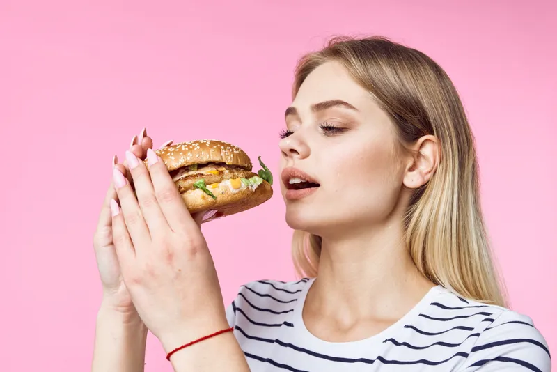 Женщина в полосатой футболке держит гамбургер — вкусный бутерброд-закуску на розовом фоне
