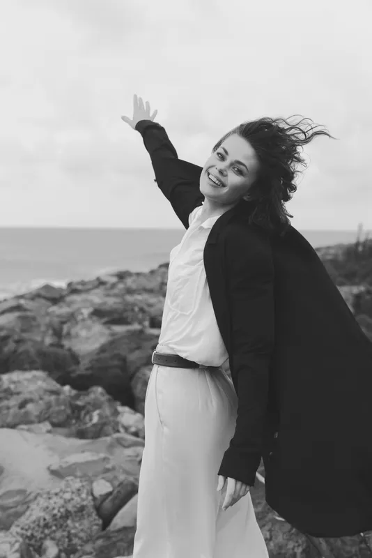 Woman on a windy beach wearing a long coat, smiling and waving, an outdoor lifestyle scene with rugged rocks and the ocean in the background.