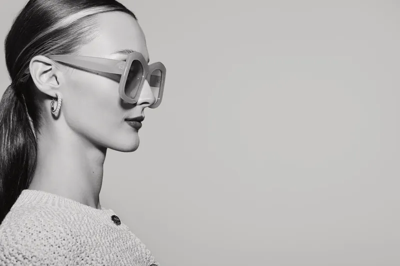 Profile of a young white woman with light skin wearing large sunglasses and a sweater on a light background.