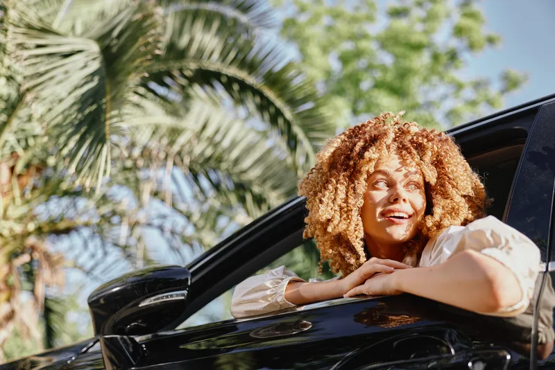 happy woman with curly hair leaning out of car window, sunny day, tropical background, natural light, casual beige shirt, smiling relaxed mood