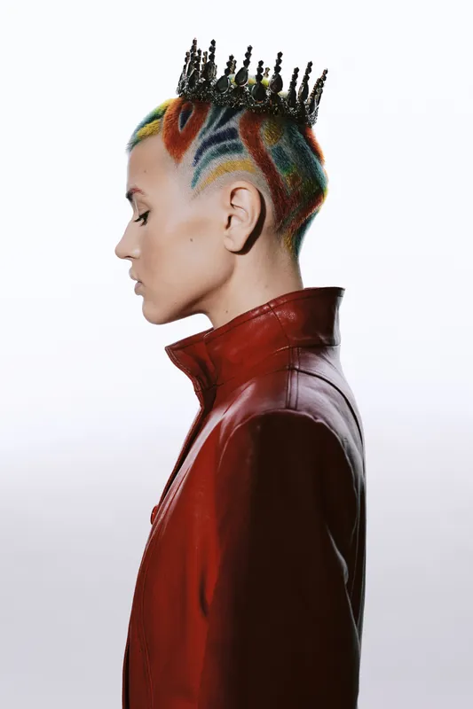 Woman with colorful patterned shaved hair wearing a red leather jacket and a decorative crown, showcasing bold fashion and unique hairstyle in profile view.
