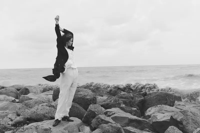 Woman standing on rocky shoreline by the sea, arms raised, wind in her jacket, black and white image capturing freedom, resilience, and outdoor adventure on a misty day.