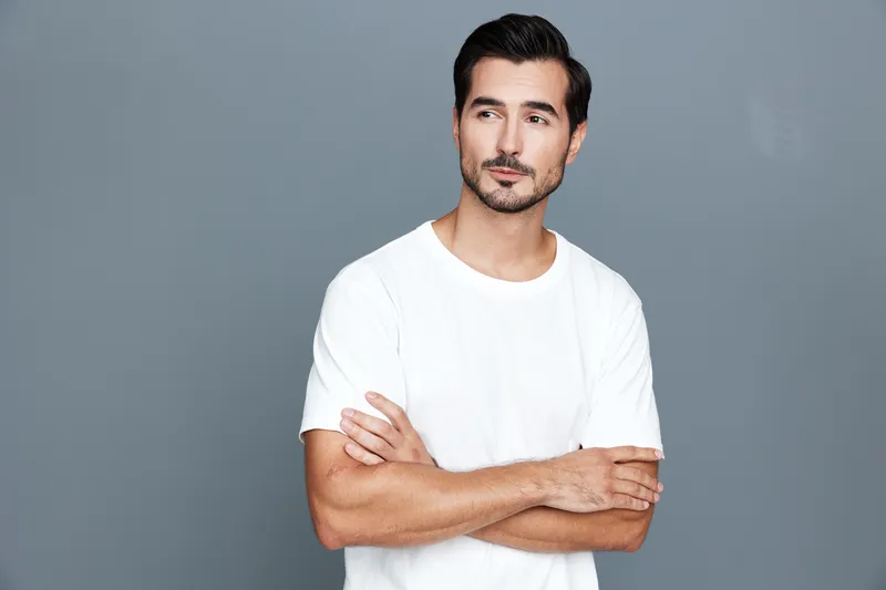 Thoughtful handsome young man with crossed arms wearing white t-shirt poses confidently against a plain gray background. Fashion and lifestyle concept.