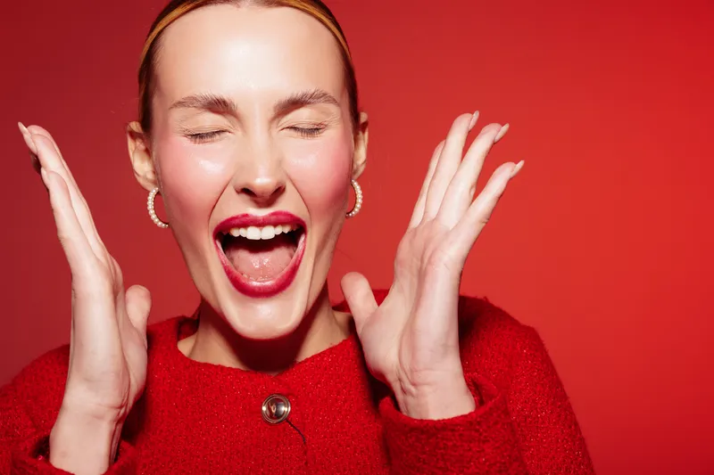 Excited woman with closed eyes and open mouth expresses joy and surprise. She wears red sweater and hoop earrings against a red background. Concept of happiness and celebration.
