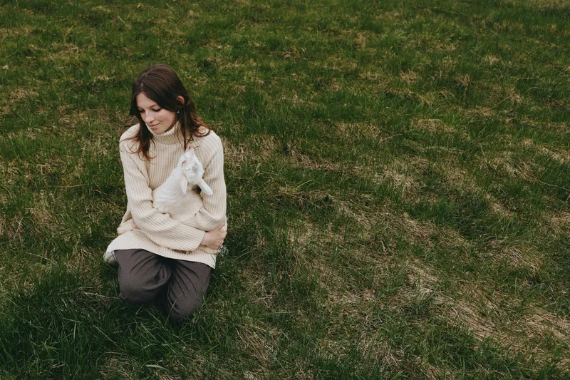 A casual lifestyle scene shows a young woman kneeling on grassy ground, cuddling a small white animal, cozy sweater, tranquil moment outdoors, conveying tenderness and connection with pets.