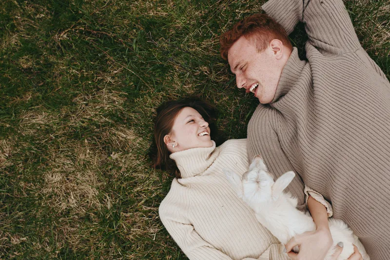 Young couple lying on grass with a baby goat cuddled in arms, wearing cozy sweaters and sharing a joyful moment outdoors in a relaxed lifestyle scene.