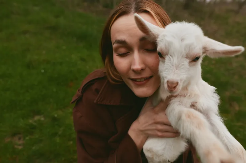 Affectionate woman warmly hugs a white goat kid outdoors, smiling softly. Natural light highlights a grassy meadow, conveying warmth, bond, and rustic lifestyle in a candid, tender moment.