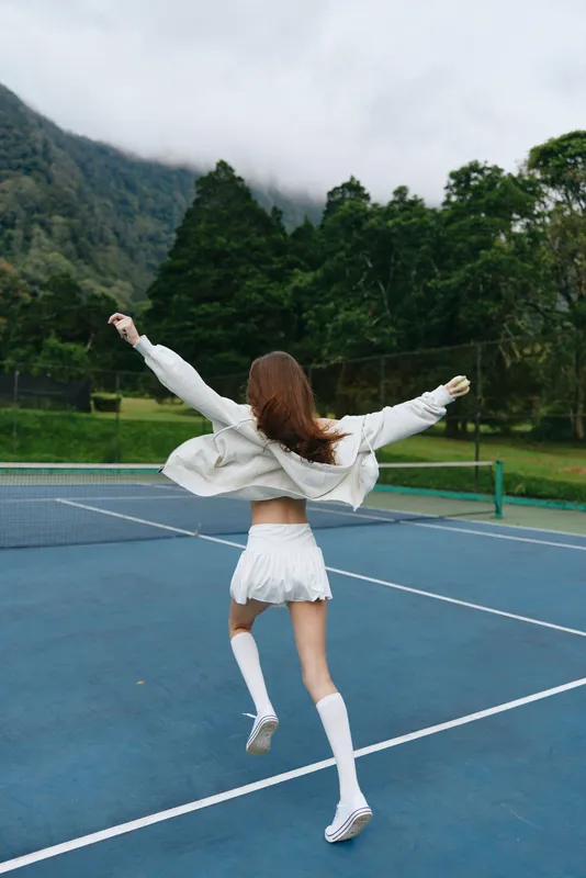 Young woman in athletic clothing joyfully running on a tennis court, showcasing energy and freedom, amidst a lush green mountain backdrop