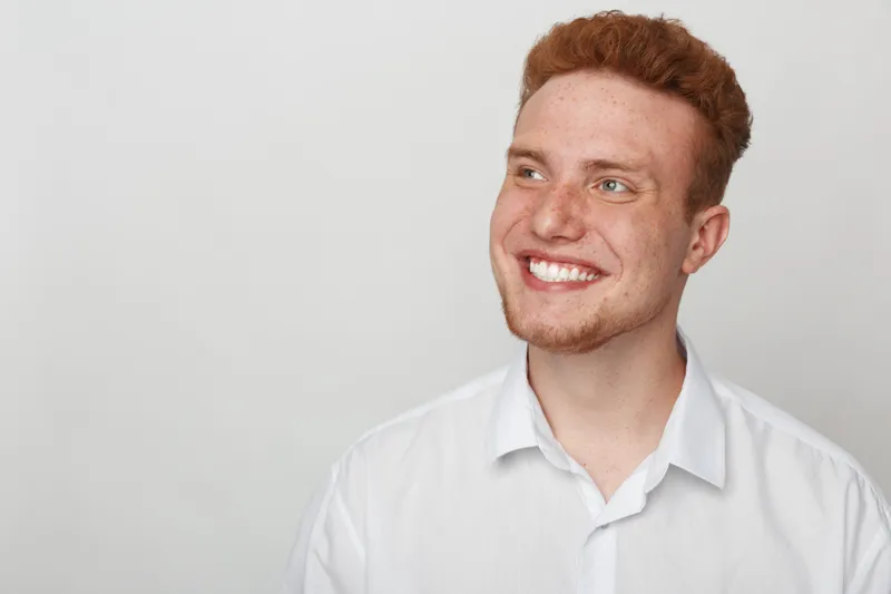 Smiling young man with red hair and beard wearing white shirt looks away with happy expression on light grey background. Concept of positive emotions, confidence, and casual portrait.