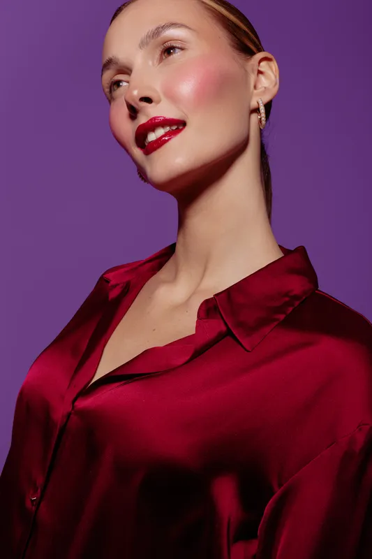 Portrait of a confident woman with red lipstick and shiny satin blouse on purple background. Studio lighting highlights her smooth skin and elegant pose. Concept of beauty and fashion.
