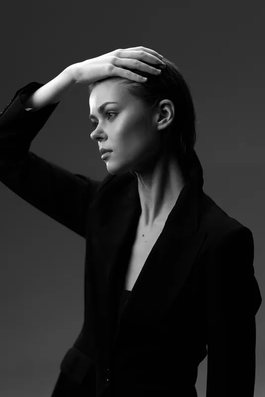 Black and white portrait of a thoughtful woman in a dark blazer with hand on forehead, expressing contemplation and elegance in a studio setting
