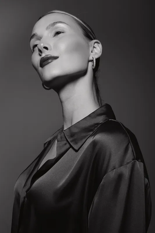 Portrait of a fair skinned caucasian woman with slicked back hair wearing shiny blouse and earrings in black and white studio lighting.