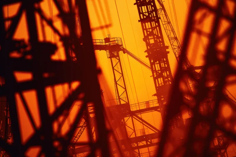 Промышленные нефтяные вышки создают яркий силуэт на фоне ярко-оранжевого заката, олицетворяя производство энергии и красоту технологий на лоне природы