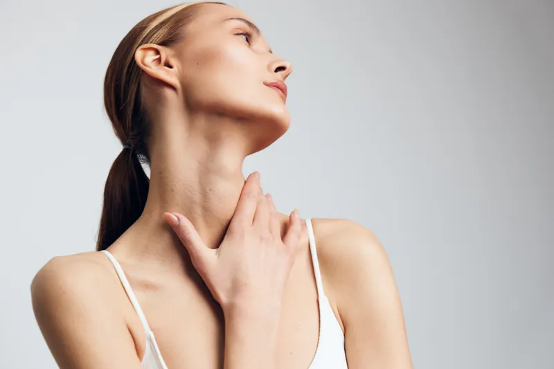 Woman skincare beauty portrait in studio lighting, she touches neck with both hands, calm relaxation expression, closeup of youthful face and collarbone against neutral background