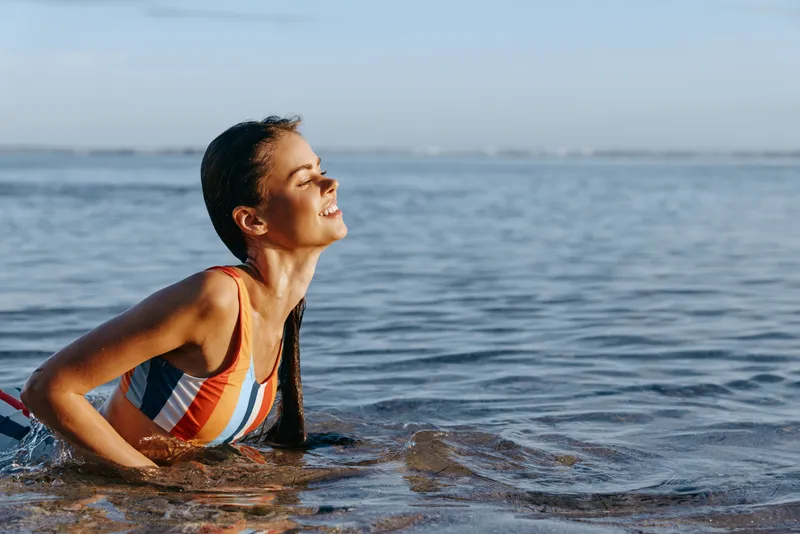 Young woman in colorful swimsuit enjoys sunny day swimming in calm sea water with relaxed and happy expression, clear sky in background during summer.