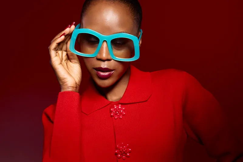 Stylish African woman with dark skin wearing bright red sweater and large blue glasses poses confidently against red background. Concept of fashion, beauty, and elegance.