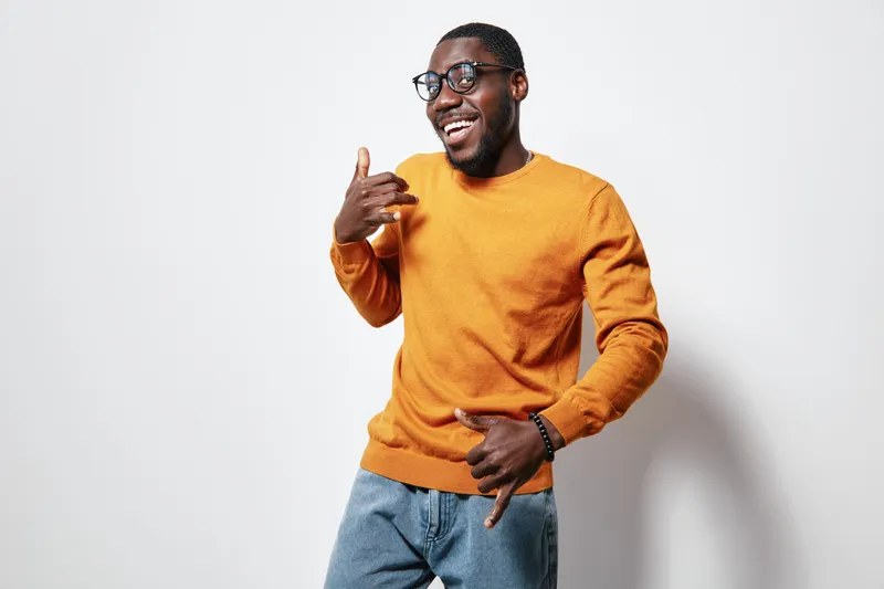 African American man with dark skin wearing glasses and orange sweater posing with hand gestures and smiling on white background in casual style.